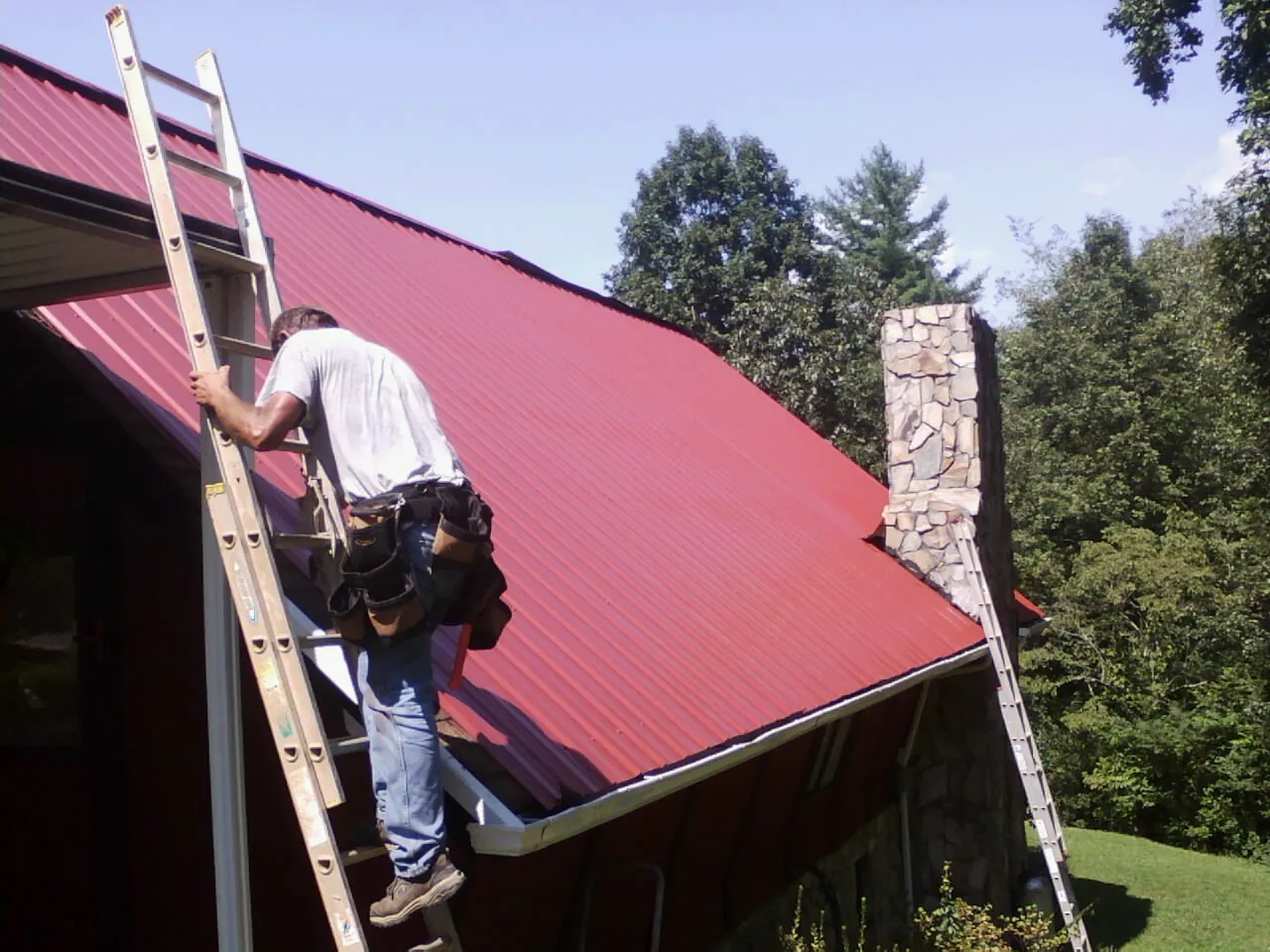 Roofer on a ladder beside a red metal roof and stone chimney — Soffit & Fascia Repair by Roofing Brothers in Pinetops